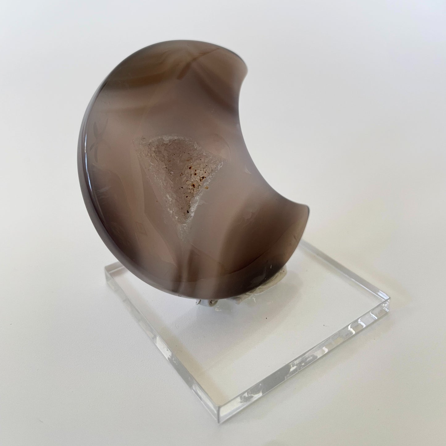 A close-up photograph of a crescent-shaped agate carving with varying shades of brown and gray. The surface is textured and features intricate patterns, with tiny druze crystals sparkling across the gray surface.