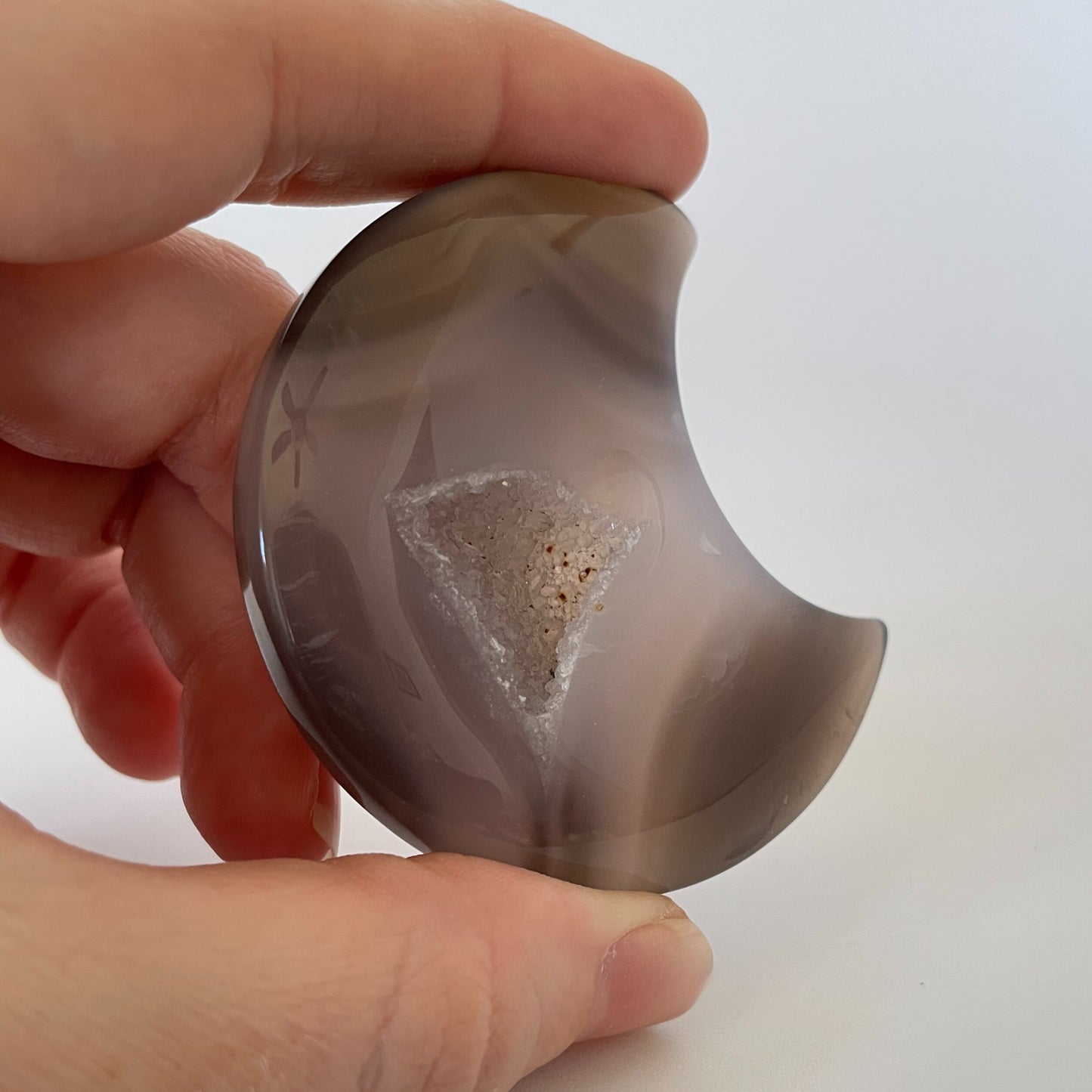 A close-up photograph of a crescent-shaped agate carving with varying shades of brown and gray. The surface is textured and features intricate patterns, with tiny druze crystals sparkling across the gray surface.