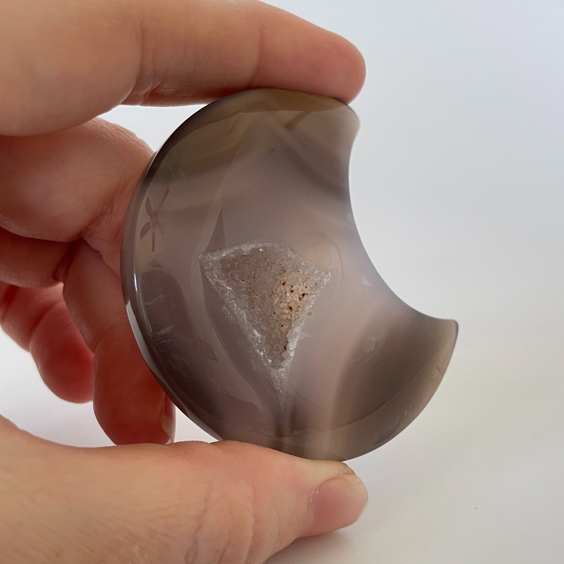A close-up photograph of a crescent-shaped agate carving with varying shades of brown and gray. The surface is textured and features intricate patterns, with tiny druze crystals sparkling across the gray surface.