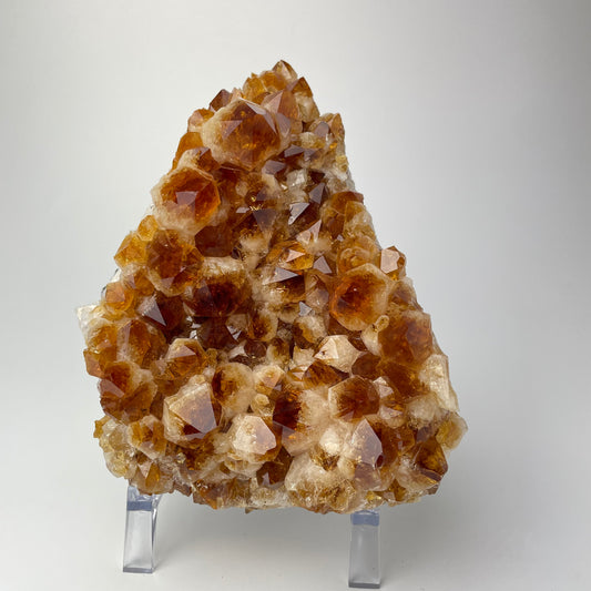 Citrine cluster with deep, orange-colored crystal formations on an acrylic stand against a gray background.
