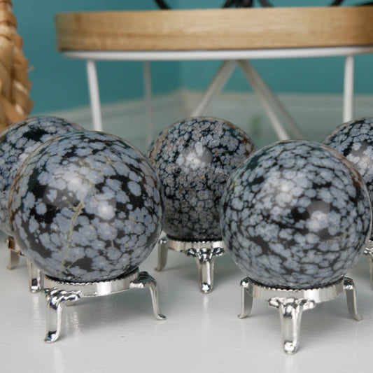 A 2-inch Snowflake Obsidian Sphere resting on a black velvet cushion. The sphere exhibits a smooth, glossy surface with unique white snowflake-like patterns dispersed across its jet-black body, creating a striking contrast. Light reflects subtly off its surface, highlighting the intricate natural formations within.