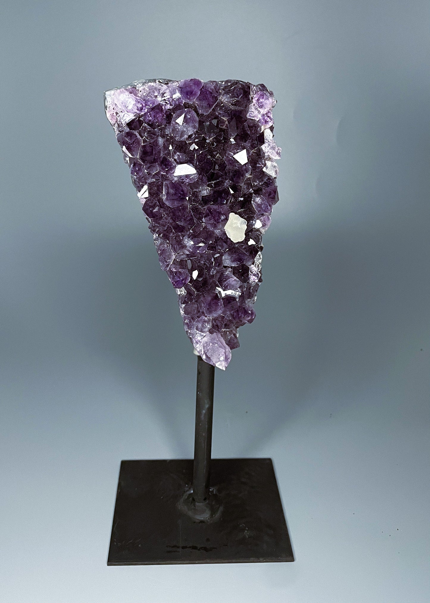 A Brazilian amethyst cluster in a triangular shape with large crystals and calcite growth on a black metal stand.