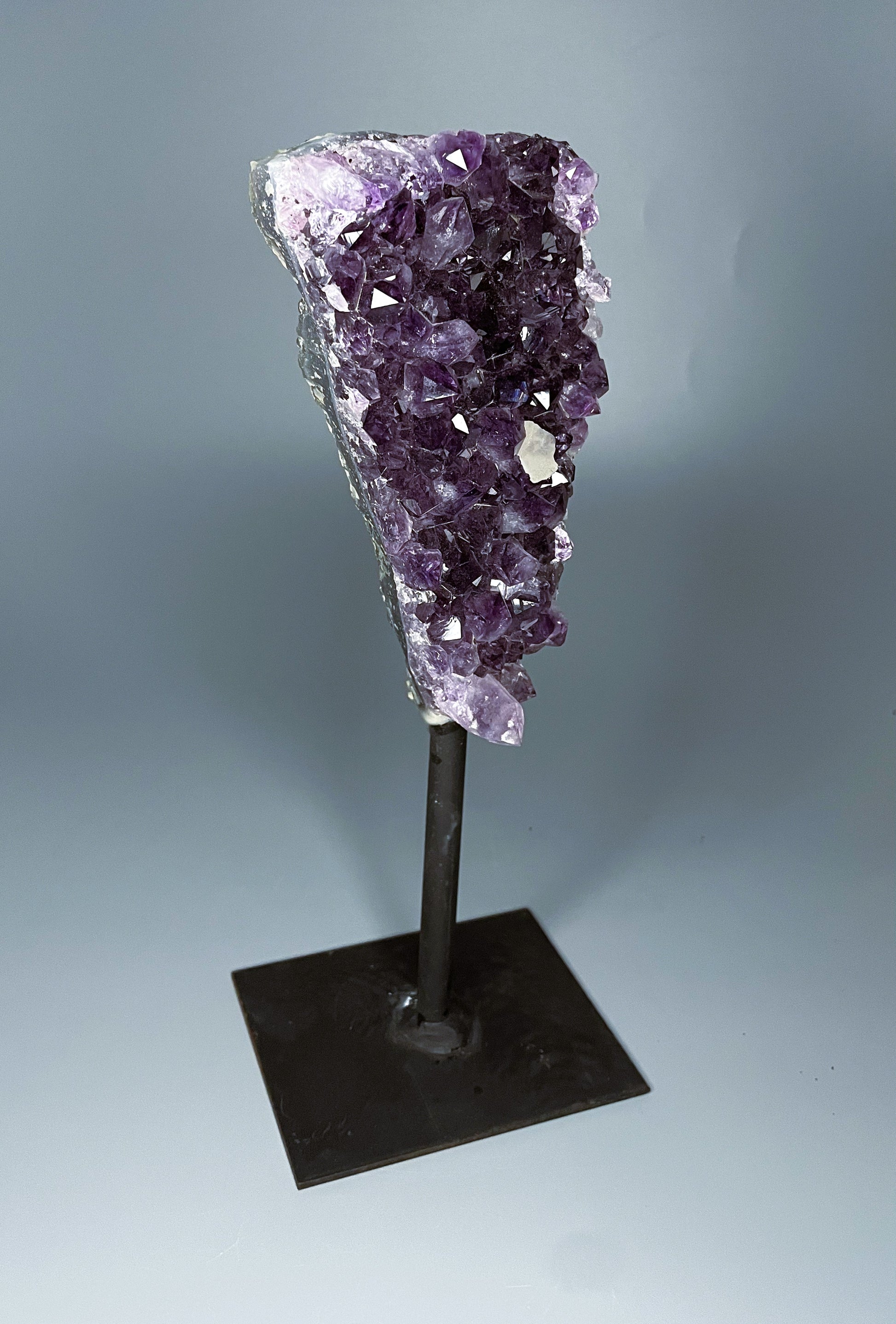 A Brazilian amethyst cluster in a triangular shape with large crystals and calcite growth on a black metal stand.