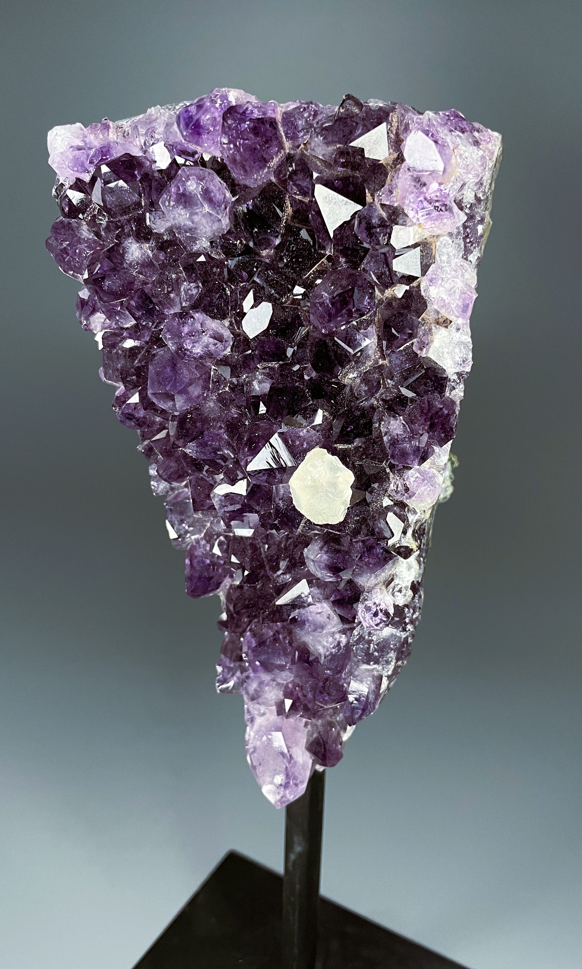 A Brazilian amethyst cluster in a triangular shape with large crystals and calcite growth on a black metal stand.