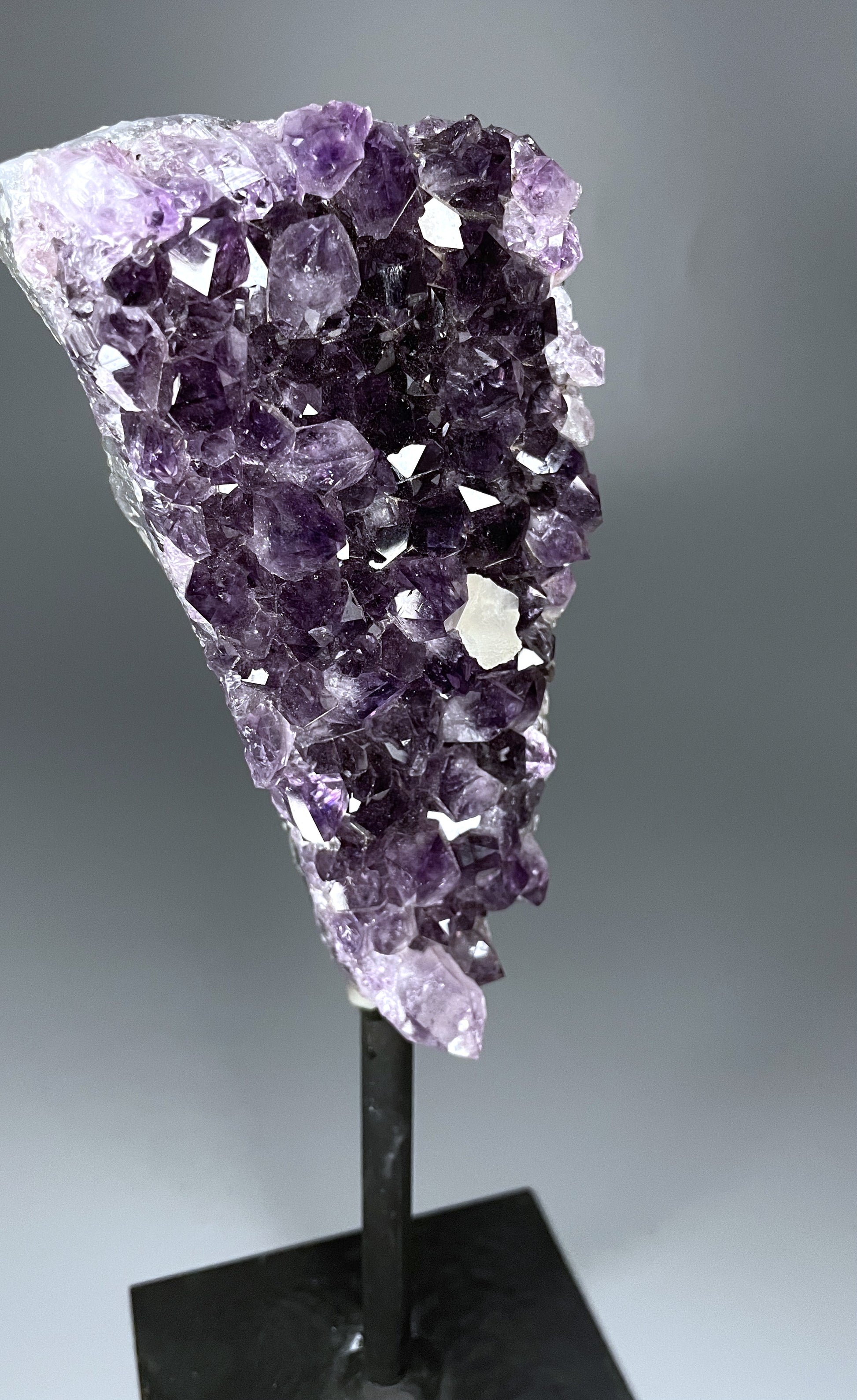 A Brazilian amethyst cluster in a triangular shape with large crystals and calcite growth on a black metal stand.