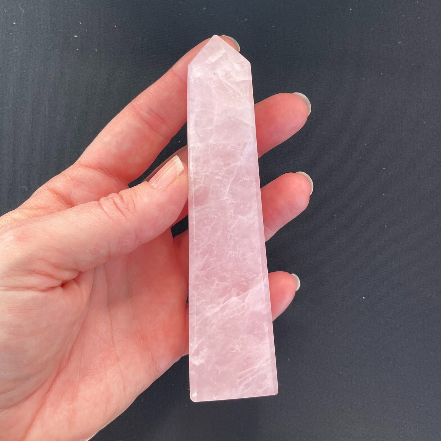 A polished rose quartz obelisk, approximately five inches in height, showcasing a soft pink hue with natural marbling and reflections of light.